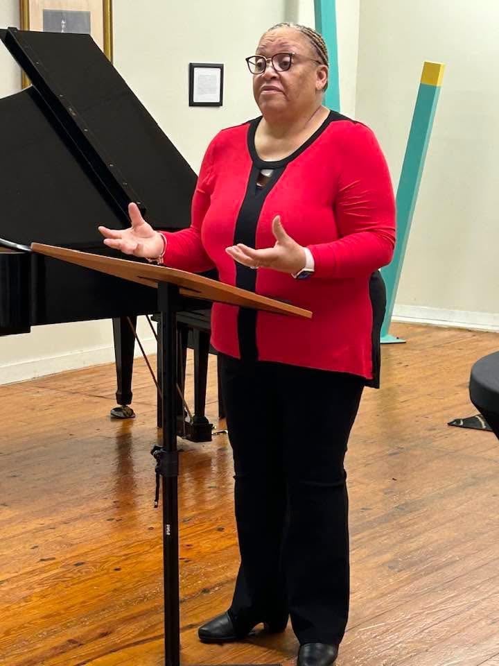 Woman in red cardigan speaking at a piano in a music studio, gesturing expressively while presenting