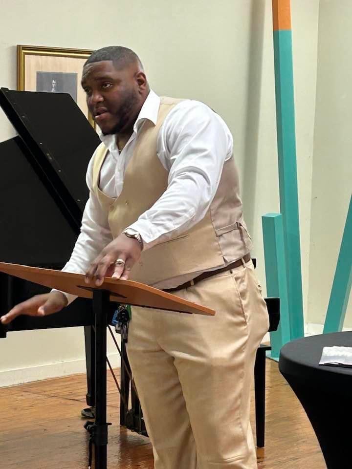 Man in beige vest and white shirt playing a black grand piano in an indoor room with pale green walls